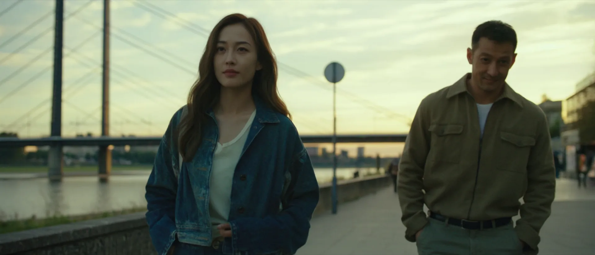 Two people in denim and a sand overshirt walking along the Rhine promenade in Düsseldorf at golden hour, the cable-stayed bridge behind them