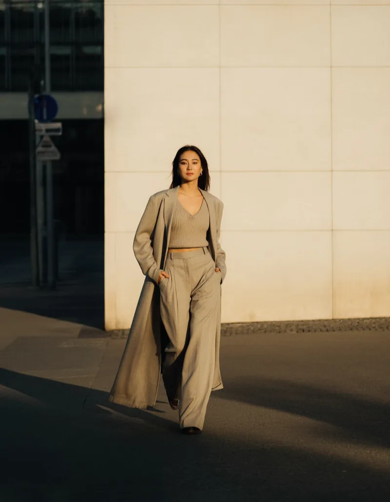 A woman in stone wide-leg trousers walking past a pale limestone wall in low evening sun