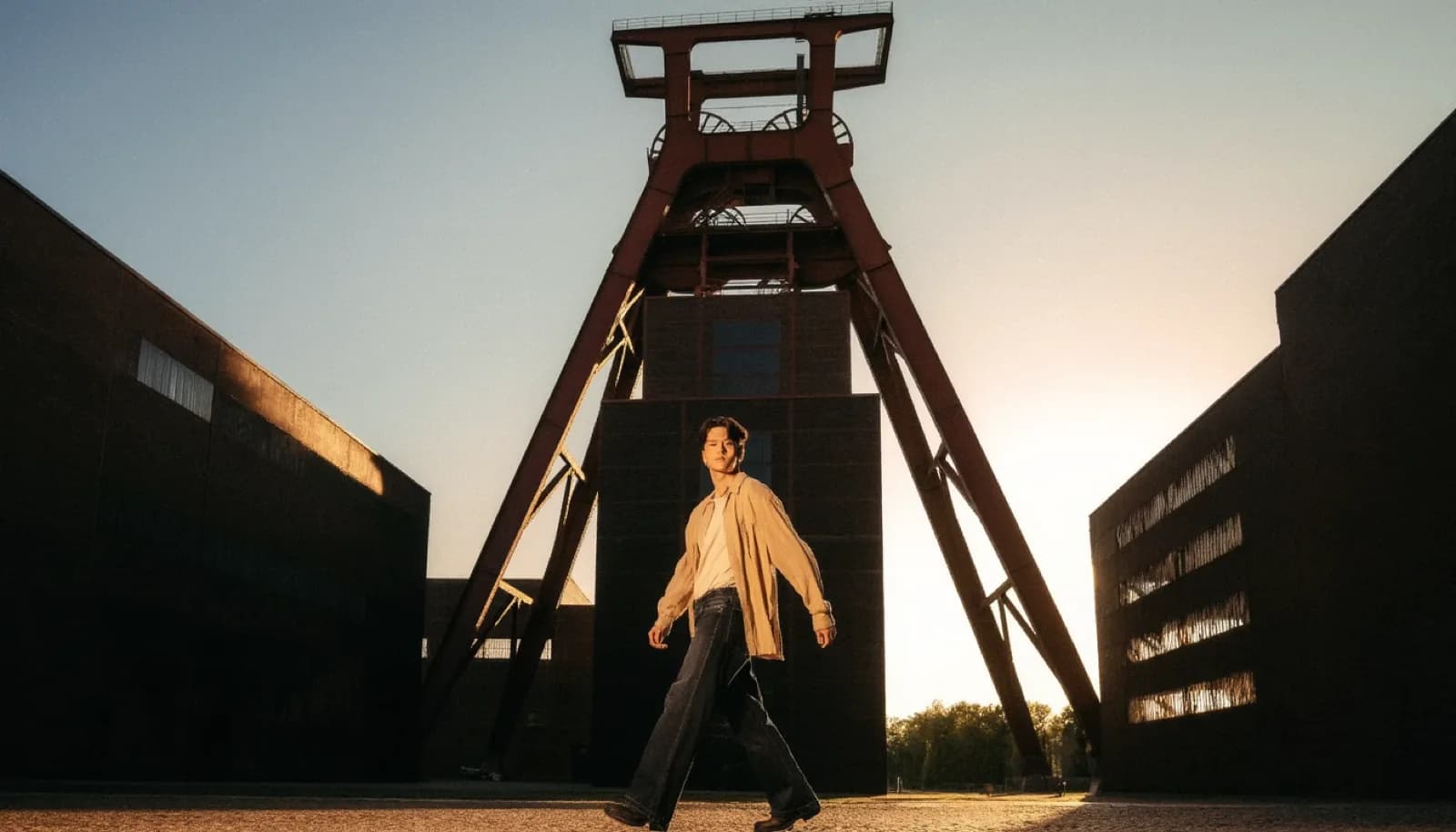 A person in a sand overshirt crossing a wide square in front of the red steel winding tower of a former Ruhr coal mine at sunset